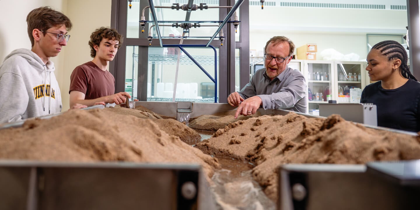 A faculty member demonstrates engineering to students at a lab table filled with dirt and water